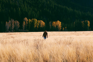 Man Walking Through Field In The Sawtooth Mountains With Mountain Range Behind Him